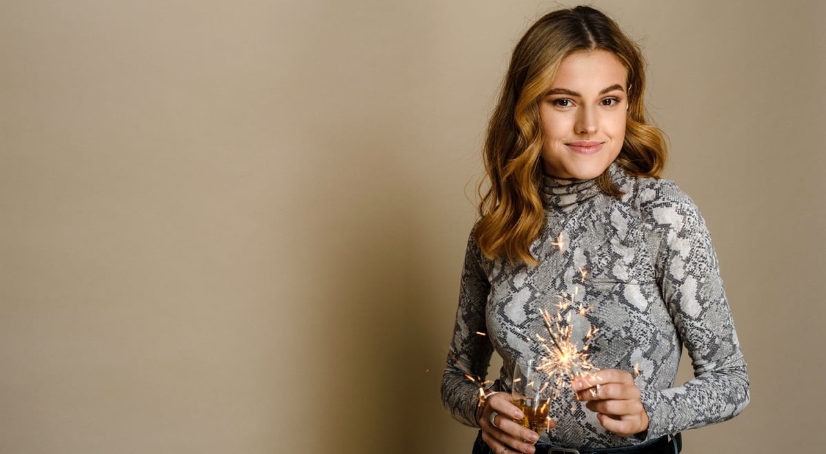 cheerful-young-woman-with-burning-sparkler-smiling-with-glass-with-champagne-sparkling-wine-beige-background